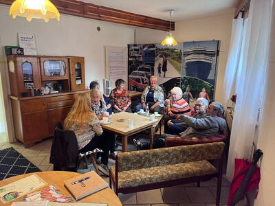 In der Wanderausstellung sitzen eine Gruppe älterer Menschen auf Sesseln an einem Tisch und unterhalten sich oder lächeln in die Kamera.