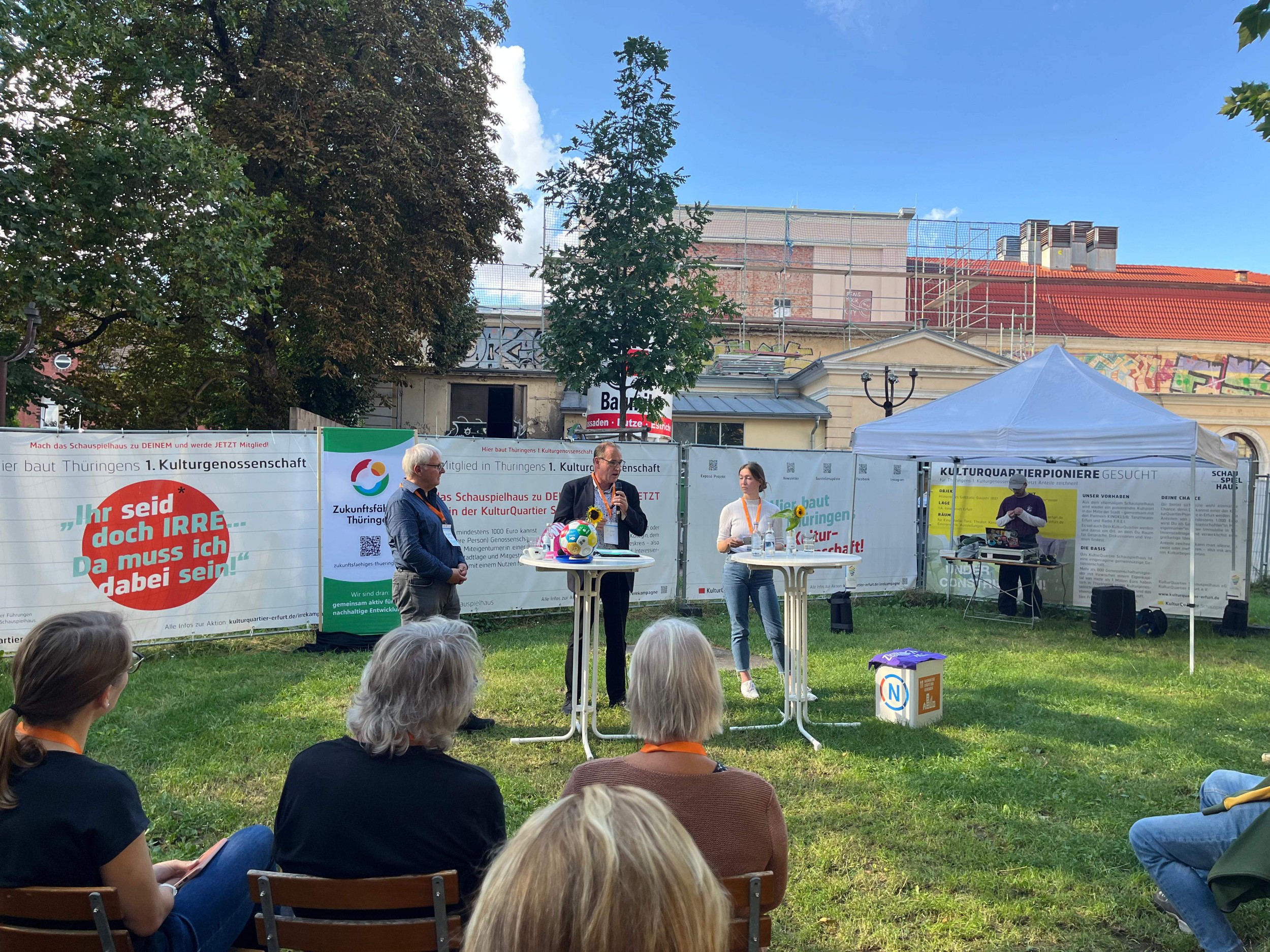 An zwei Stehtischen stehen drei Personen, wovon eine Person in ein Mikro spricht. Vorne sind abgeschnitten einige Menschen zu sehen, die sitzend der Rede zuhören. Im Hintergrund sind ein Pavillon und mehrere gestaltete Stoffwände zu sehen.